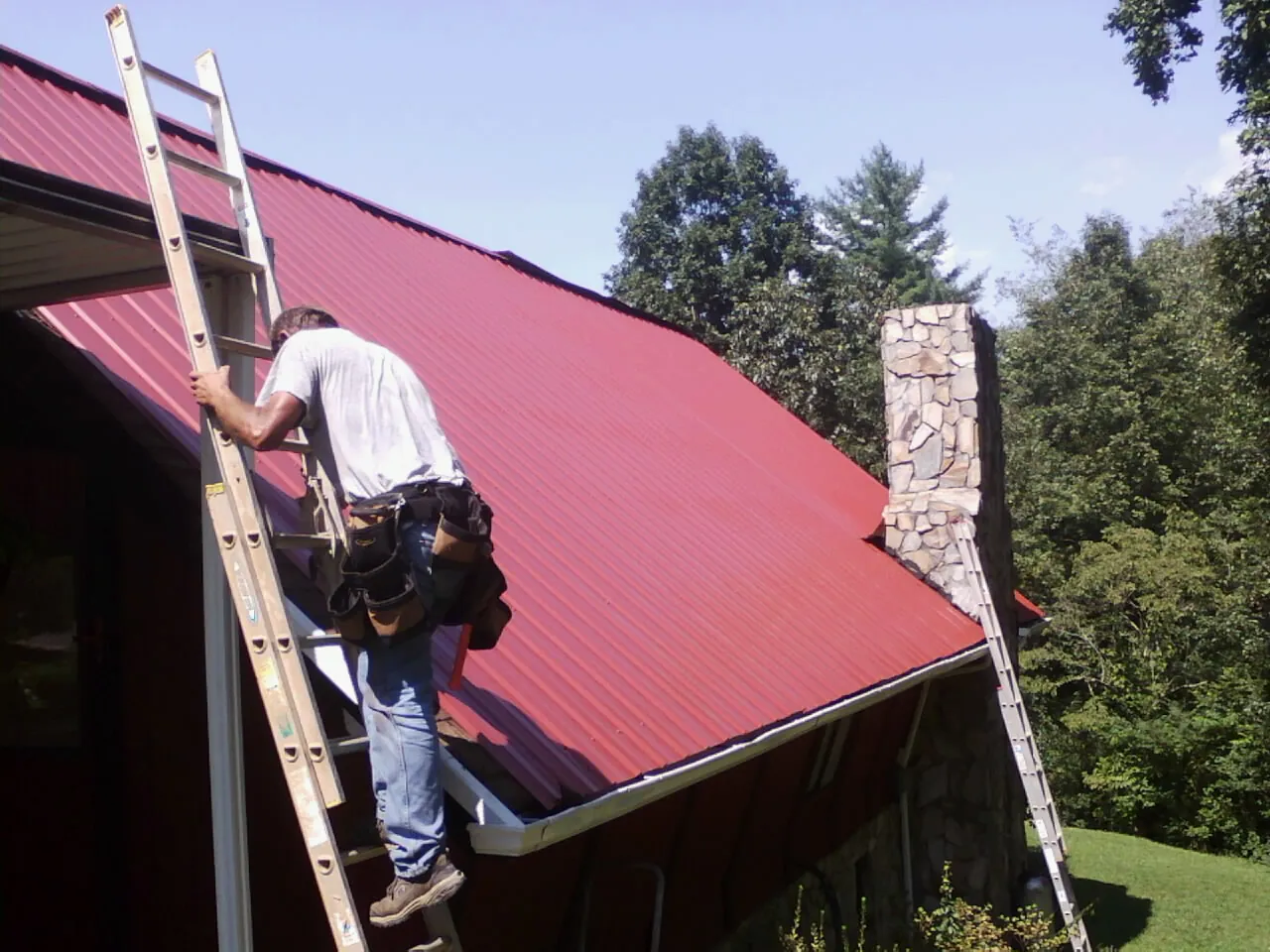 Roofer on a ladder beside a red metal roof and stone chimney — Soffit & Fascia Repair by Roofing Brothers in La Canada Flintridge